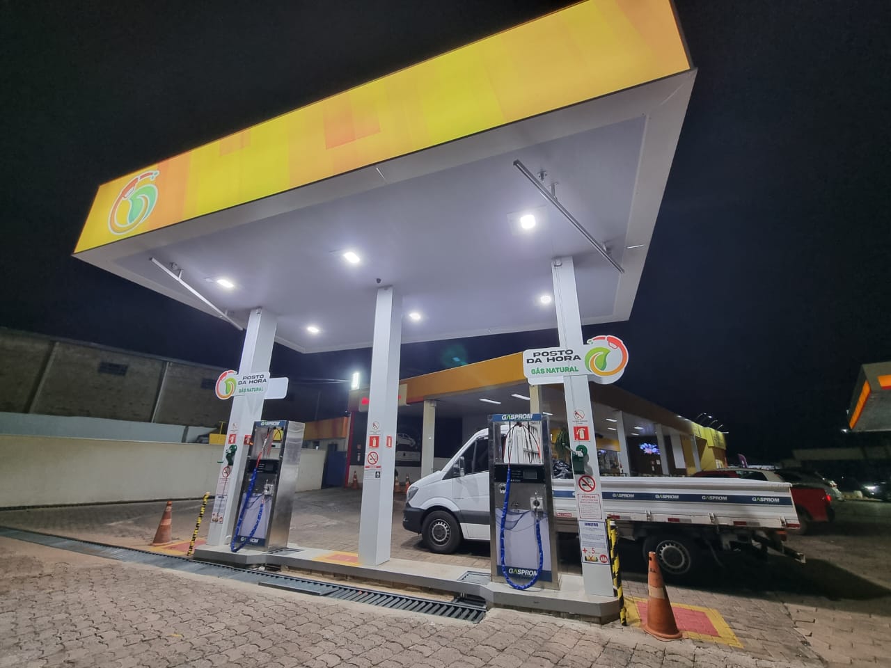 Pista de posto de GNV iluminada à noite com dois dispensers Gasprom e caminhão com carroceria Gasprom